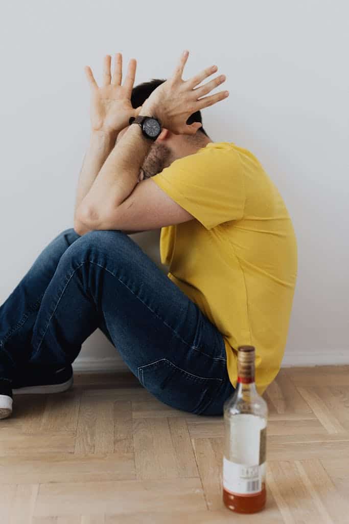 A man in emotional distress sits indoors with an alcohol bottle symbolizing mental health struggles.