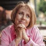 Senior woman with short brunette hair and pink shirt, hands clasped, looking thoughtful outdoors.